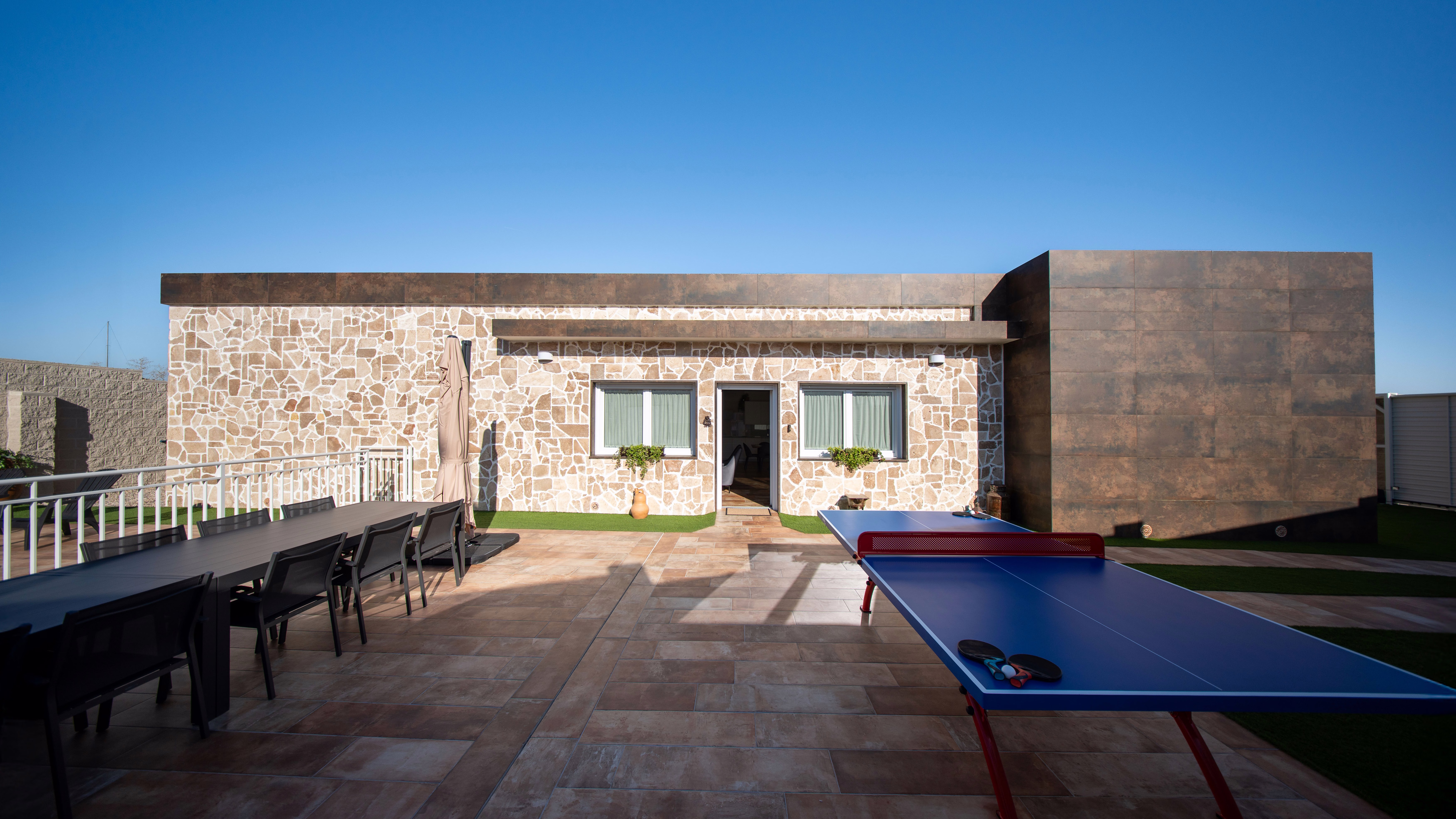 Amplia terraza con mesa de ping‑pong, larga mesa de comedor al aire libre y fachada de piedra; un espacio soleado ideal para reuniones y ocio.
